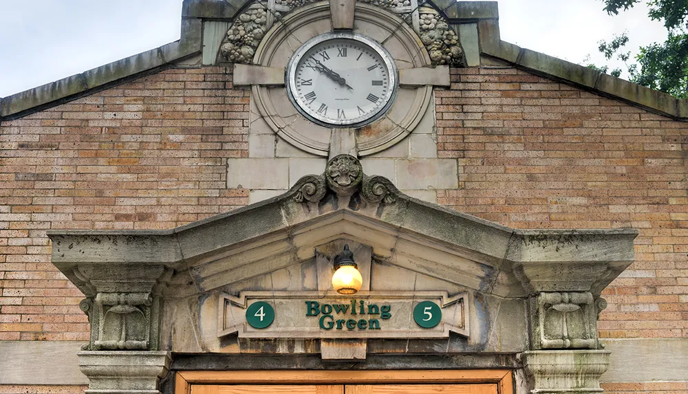 The image shows the classic facade of the Bowling Green subway station with its ornate stonework clock and a lit lamp indicating a historic setting