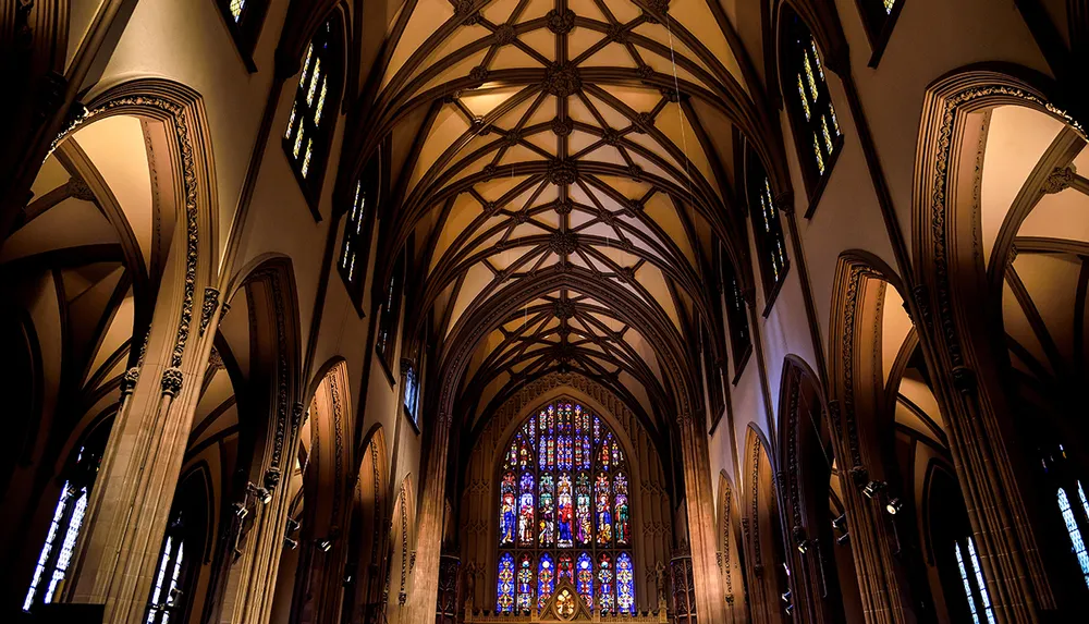 The image shows the intricate Gothic architecture and beautifully colored stained glass windows of a cathedrals interior with a focus on its ribbed vaulting and arched designs