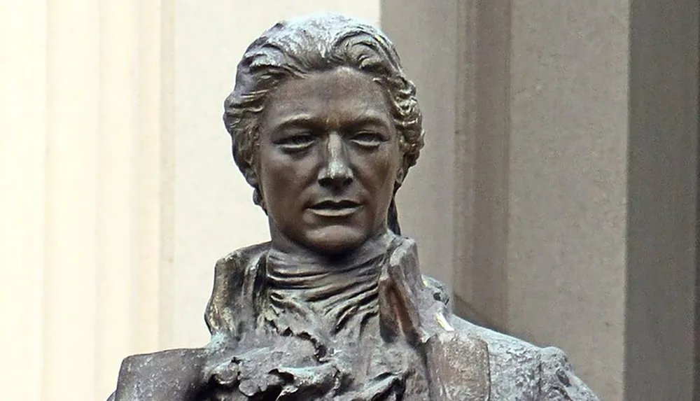 The image shows a close-up of a bronze statue of a historical figure set against a blurred background with architectural elements