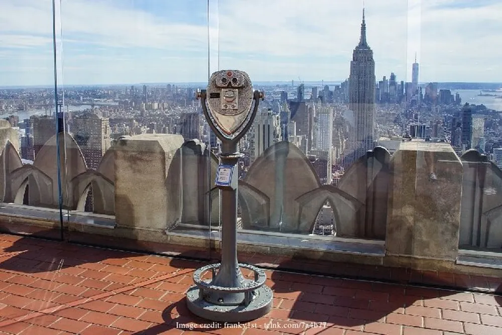 A coin-operated binocular viewer stands on an observation deck with a clear view of a city skyline featuring a prominent skyscraper under a blue sky