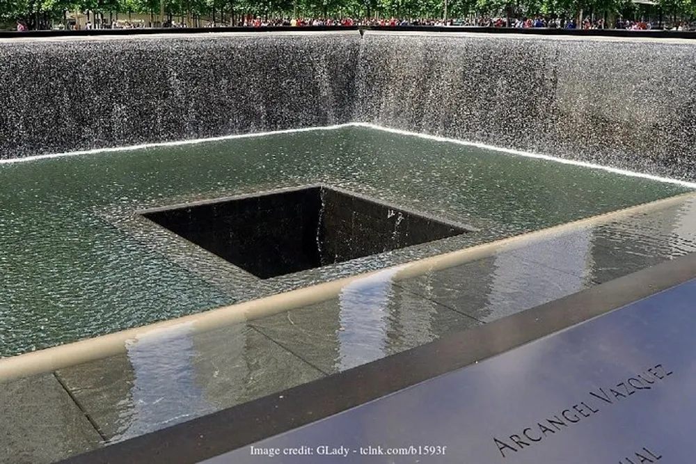 The image shows one of the Reflecting Pools at the 911 Memorial in New York City designed as a tranquil and contemplative space with water cascading down the sides and disappearing into a central void