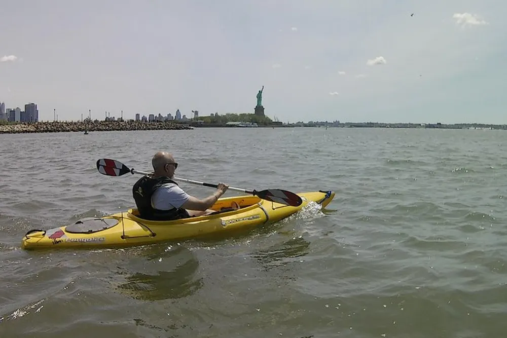 A person is kayaking on the water with the Statue of Liberty and New York skyline in the background