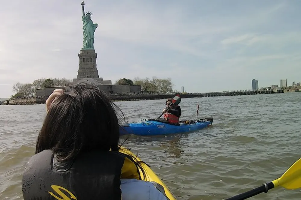 A person in a kayak is paddling near the Statue of Liberty observed by another individual who seems to be in a kayak as well