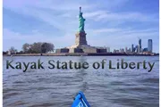 The image shows the view from a kayak on the water near the Statue of Liberty with the New York City skyline in the background.