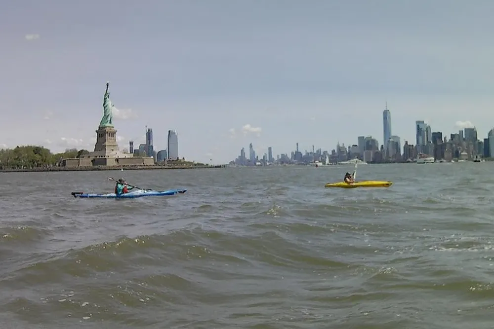 Two kayakers are paddling near the Statue of Liberty with the New York City skyline in the background