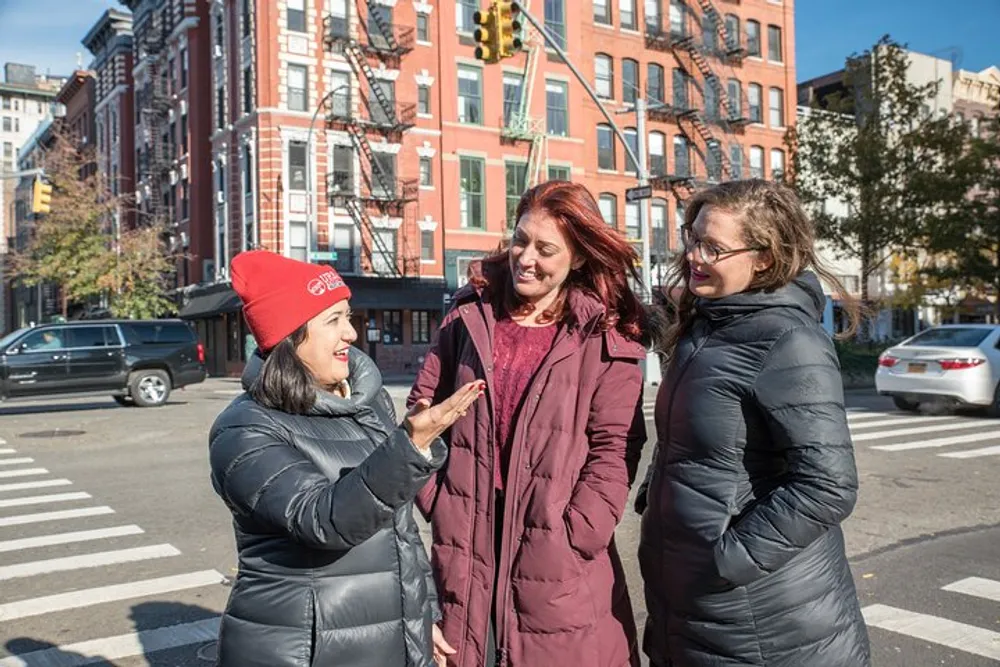 Three women are engaged in a friendly conversation on a sunny city street corner with one of them gesturing expressively