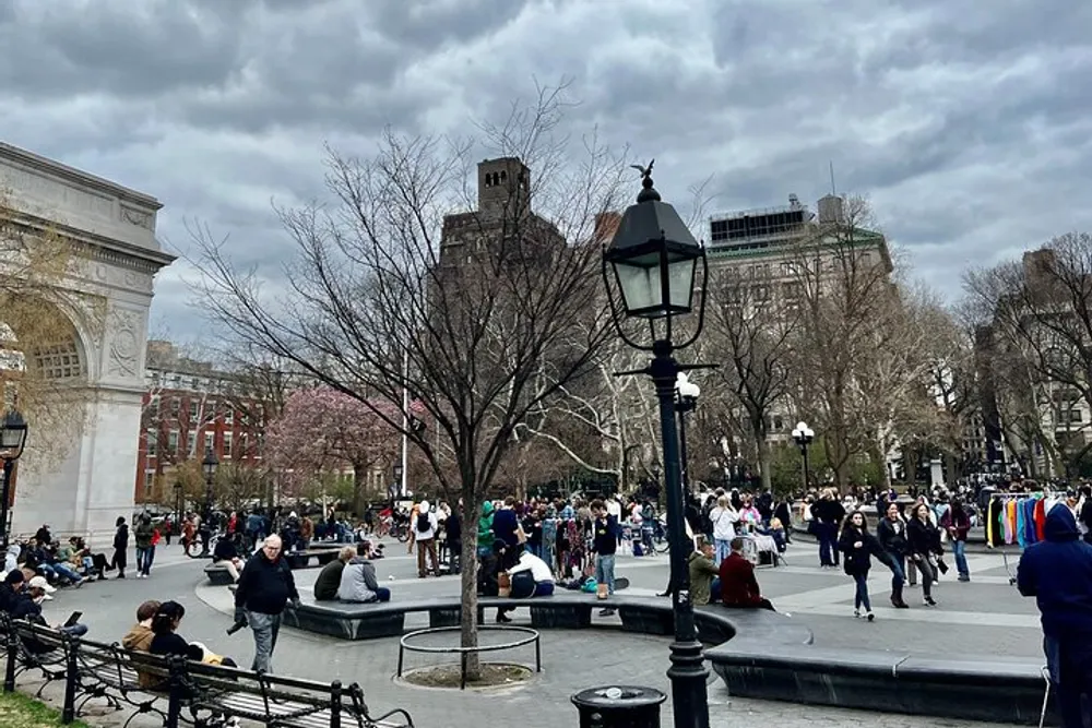 The image shows a bustling urban park with people walking around sitting on benches and enjoying an overcast day with a prominent arch structure in the background