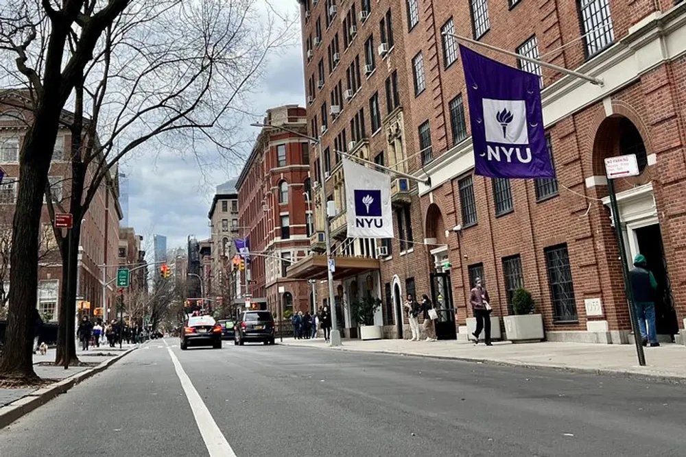 The image shows an urban street scene with NYU flags on a building indicating it is a part of New York University with pedestrians and vehicles and a view of distant skyscrapers