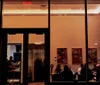 The image captures a warm and inviting view into a restaurant at night seen from the outside through large windows where people are dining and conversing under the soft interior lighting with colorful artwork on the walls