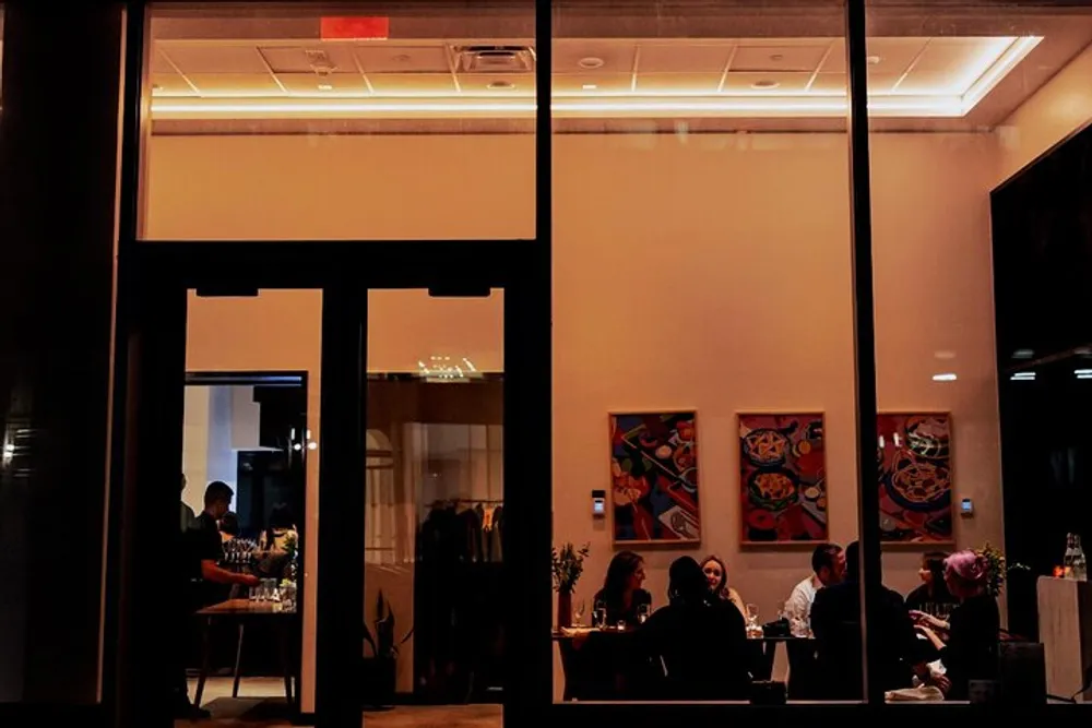 The image captures a warm and inviting view into a restaurant at night seen from the outside through large windows where people are dining and conversing under the soft interior lighting with colorful artwork on the walls