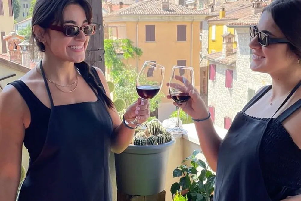 Two people are smiling and toasting with glasses of red wine on a balcony with a picturesque view of terracotta roofed buildings in the background