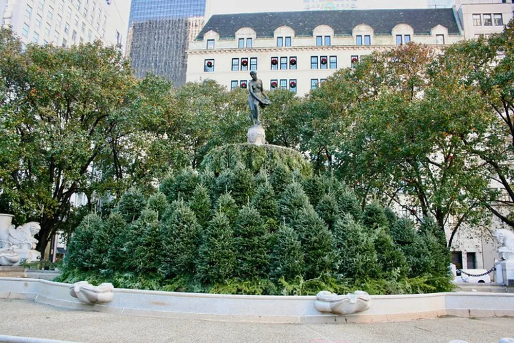 The image features a bronze statue atop a tiered tree-lined fountain in an urban park setting with city buildings in the background