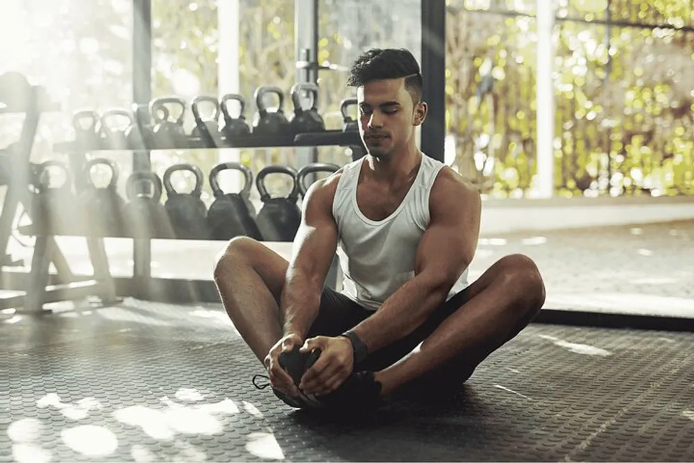 A person is stretching on the floor of a gym with weights in the background bathed in natural light coming through large windows