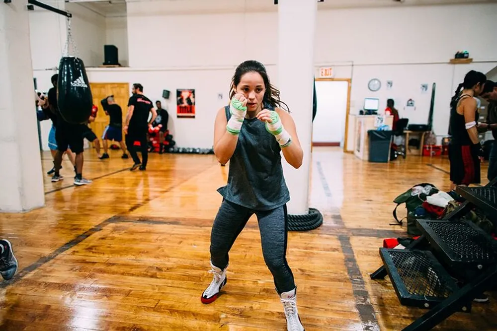 A person with boxing gloves is practicing or posing in a boxing stance in a gym with other individuals training in the background