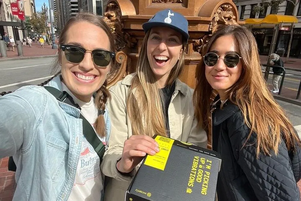 Three smiling individuals are taking a selfie on a sunny day on a city street holding a black binder or folder with a bright yellow label