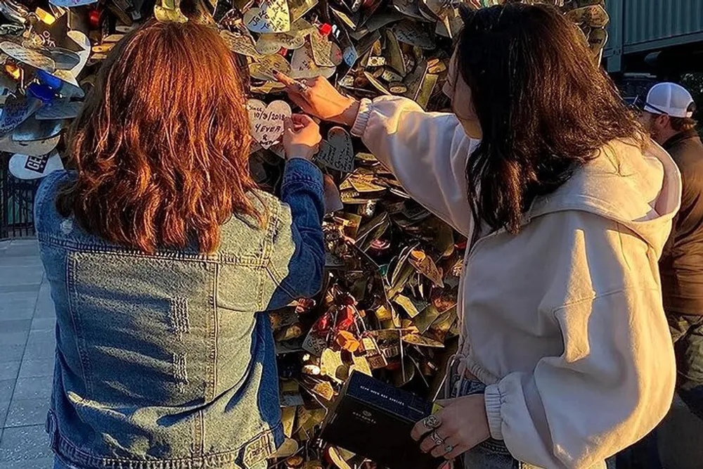 Two individuals are attaching a lock to a collection of many other locks on what appears to be a love lock fence a common symbol of commitment and affection between couples