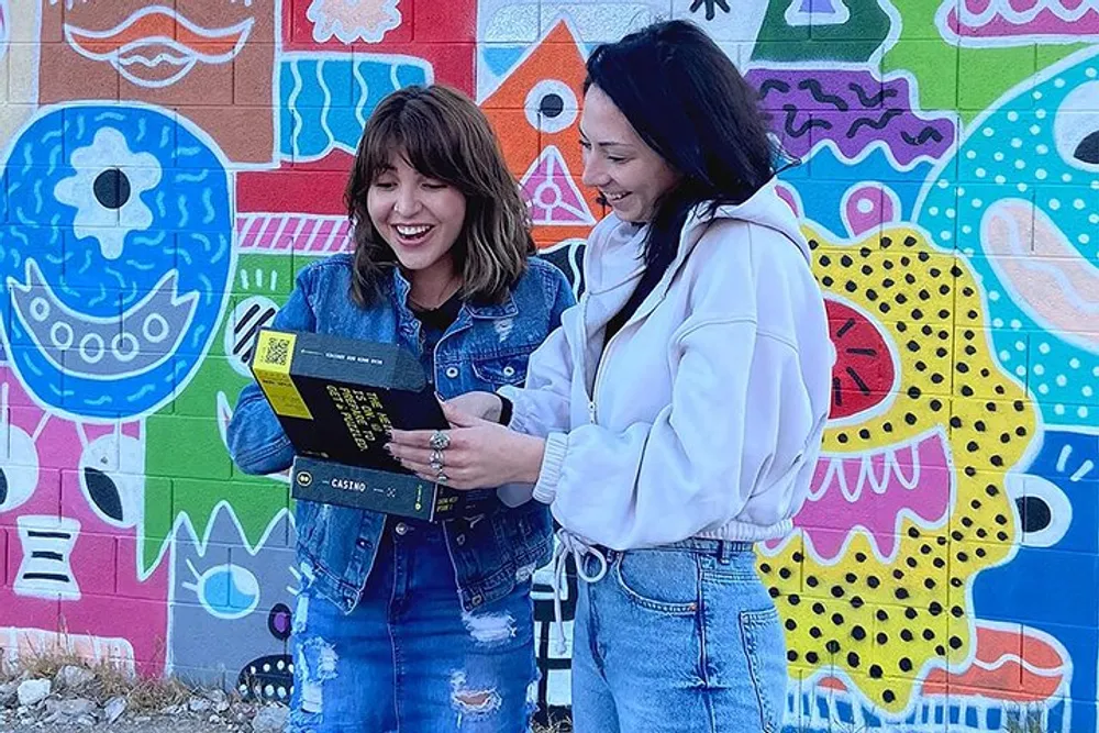 Two people are smiling and looking at a book together in front of a colorful mural wall