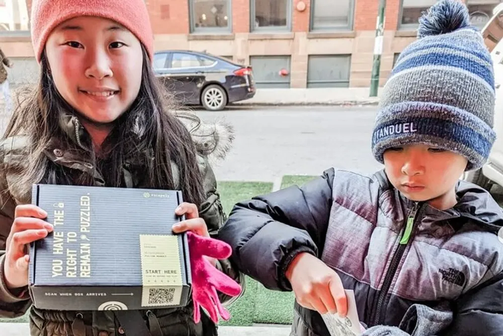 Two children are standing outdoors the girl on the left is smiling and holding a puzzle box while the boy on the right appears to be examining something in his hand with a focused expression against an urban backdrop