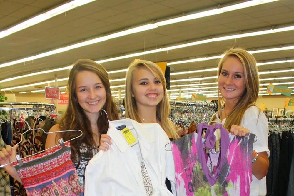 Three people are smiling and holding up clothes hangers with various items of clothing in a retail store environment