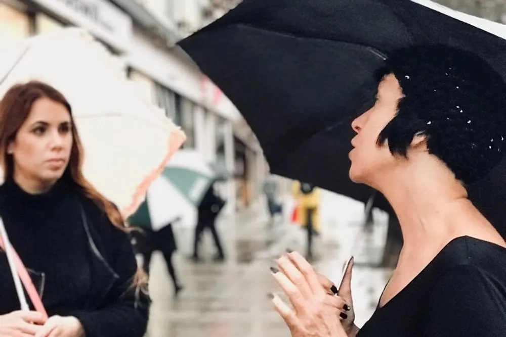 A woman with a black sequined beret is holding an umbrella and appears to be talking while another woman with long brown hair looks on against a backdrop of people with umbrellas on a rainy day