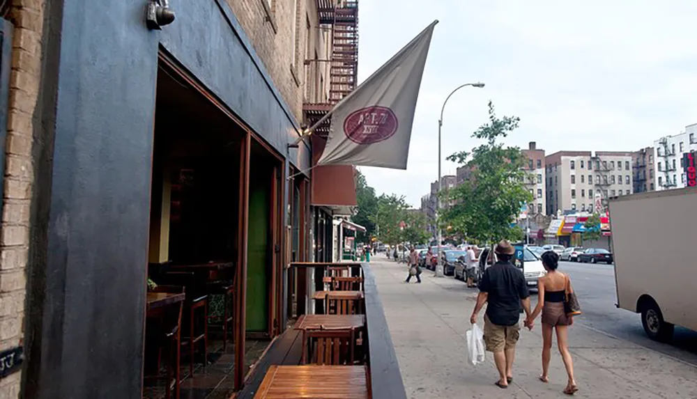 A couple walks hand in hand down a bustling city sidewalk past a caf with outdoor wooden tables and a flag bearing a logo