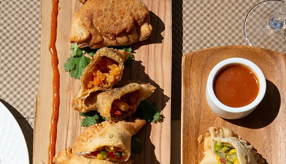 The image shows several golden-brown empanadas on a wooden serving board with a side of red sauce with one empanada cut open to reveal the colorful vegetable filling