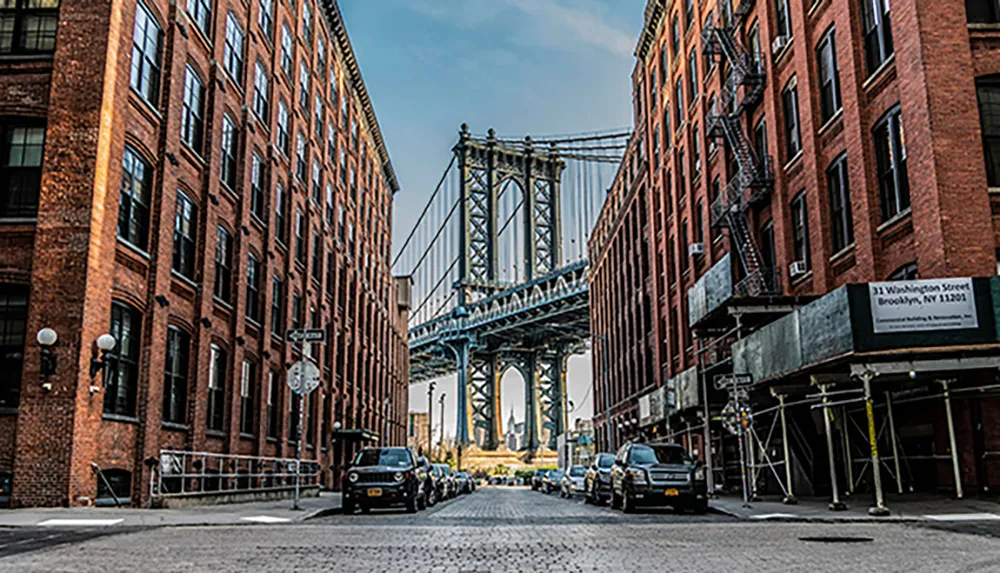 This is an iconic view down a street in DUMBO Brooklyn showcasing the Manhattan Bridge framed between old brick buildings under a clear sky