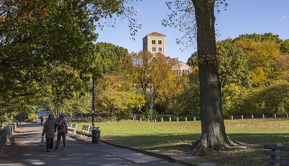 A couple walks hand in hand along a tree-lined pathway in a park during autumn with a historical tower building in the background