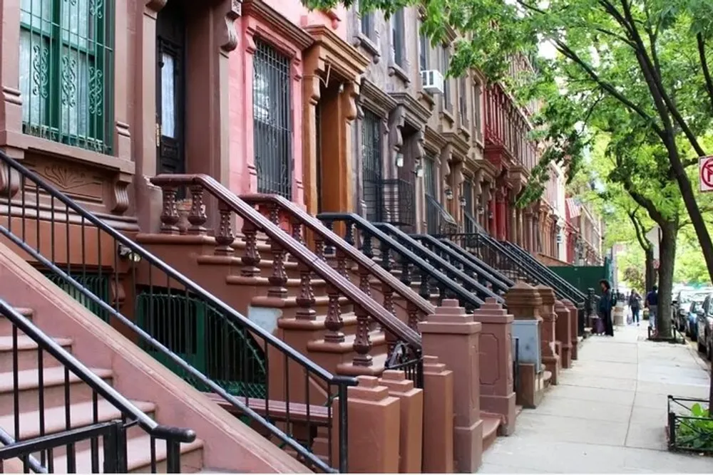 The image shows a tree-lined residential street with a row of brownstone townhouses characterized by their uniform stoops and traditional architecture