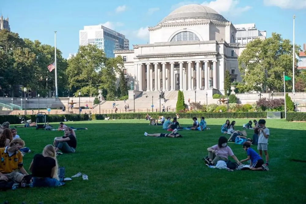 Groups of people are relaxing and socializing on a grassy lawn in front of an imposing building with a large dome and columns indicative of a university or governmental structure on a sunny day with scattered clouds