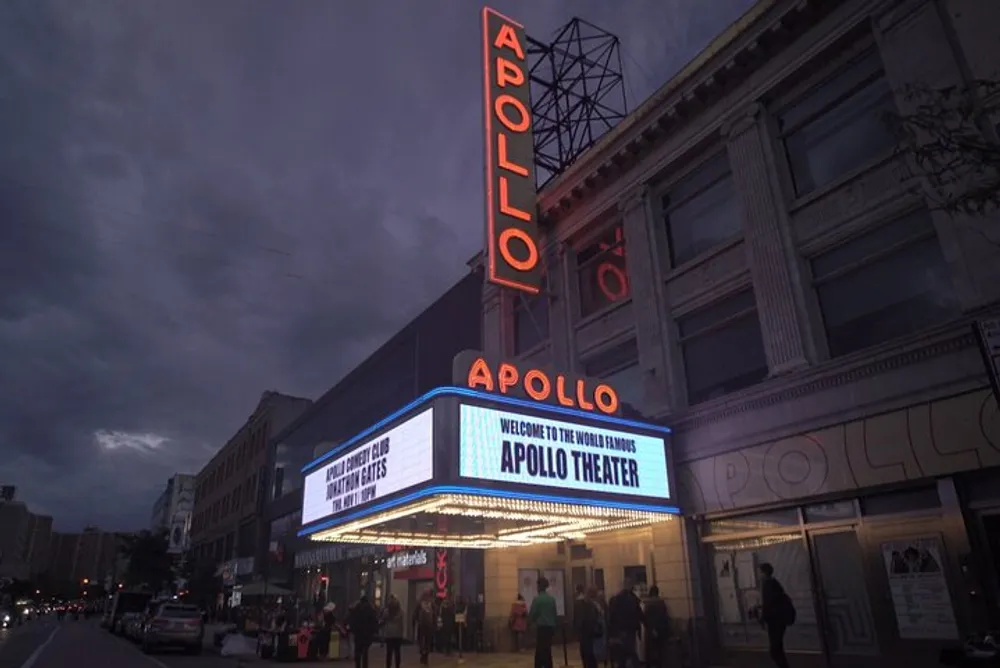 A photo of the Apollo Theater at dusk with its neon signs illuminated and people gathered outside