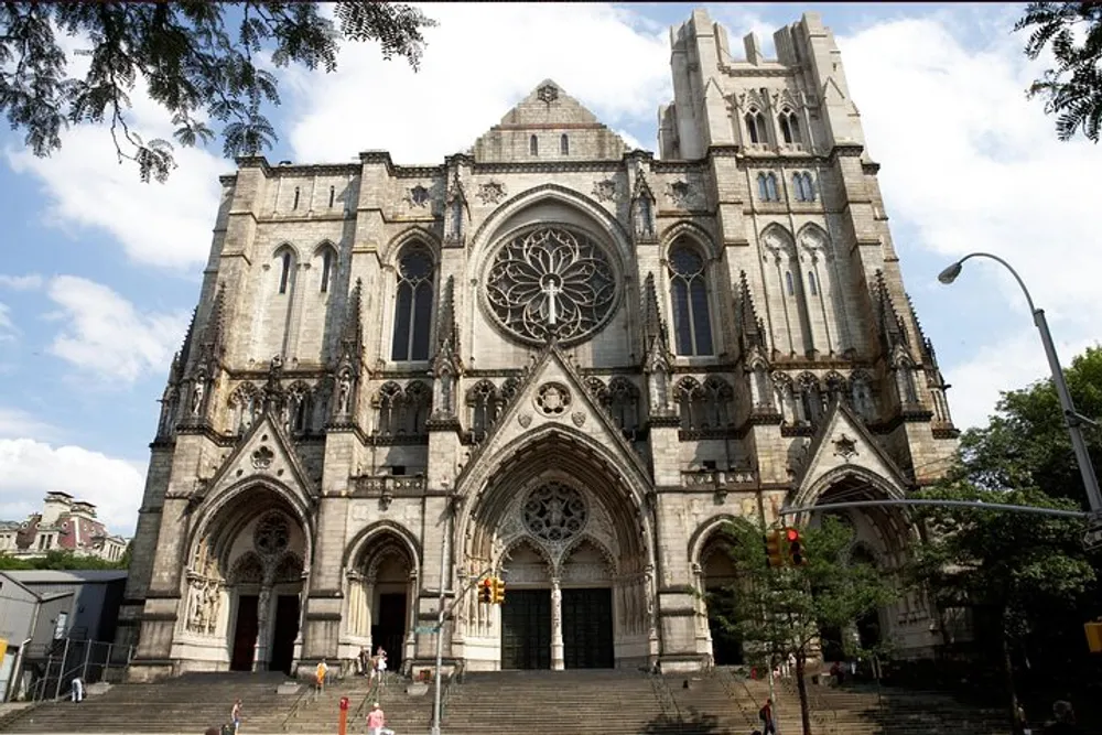 The image shows a grand gothic-style cathedral with a large rose window and several people walking by its front steps on a sunny day with partial cloud cover