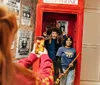 A person is taking a photo of two smiling individuals playfully posing with props inside a British-style red telephone box adorned with Harry Potter-themed decorations