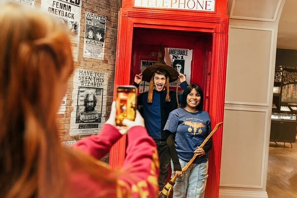 A person is taking a photo of two smiling individuals playfully posing with props inside a British-style red telephone box adorned with Harry Potter-themed decorations