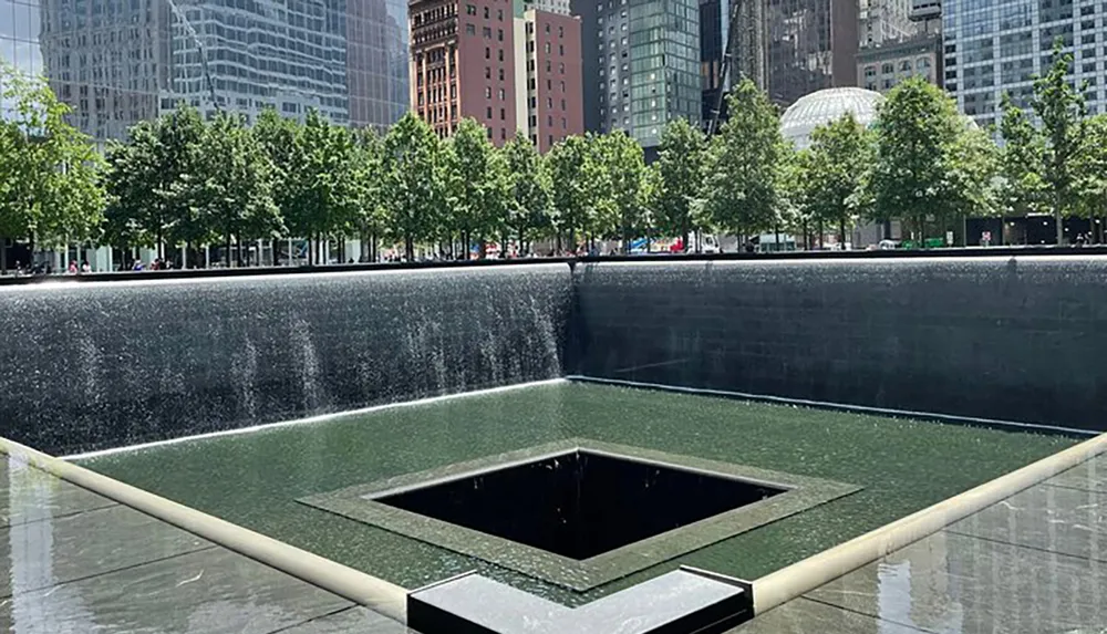 The image shows a serene memorial water feature in an urban park setting with surrounding skyscrapers and green trees under a clear sky