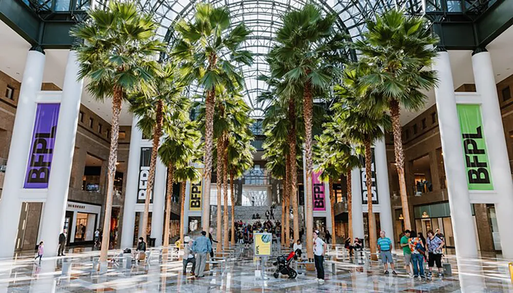 The image shows an indoor atrium with tall palm trees visitors and shops beneath a large glass dome