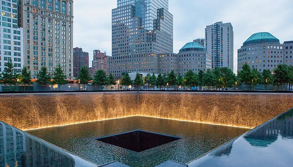 This image shows a memorial reflecting pool surrounded by trees and skyscrapers in an urban setting at twilight