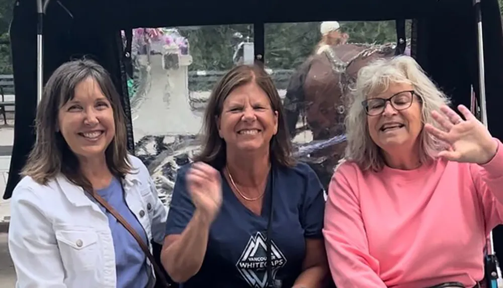 Three women are smiling and waving at the camera seemingly enjoying a ride in a vehicle with a reflective surface behind them displaying distorted reflections