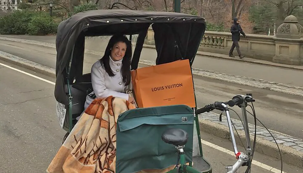 A smiling individual is seated comfortably under a blanket in a pedicab with a prominent Louis Vuitton shopping bag next to them