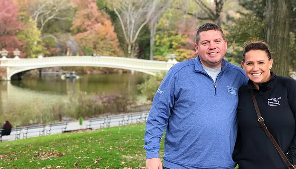 A smiling man and woman pose for a photo in front of a scenic bridge over a pond in a lush park