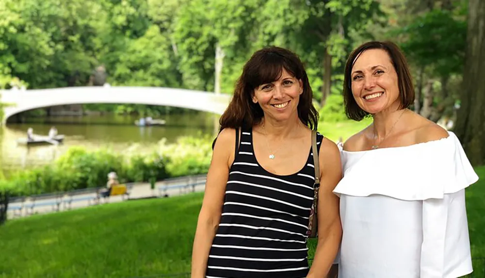 Two smiling women pose for a photo in front of a scenic pond with a white bridge and rowboats in the background