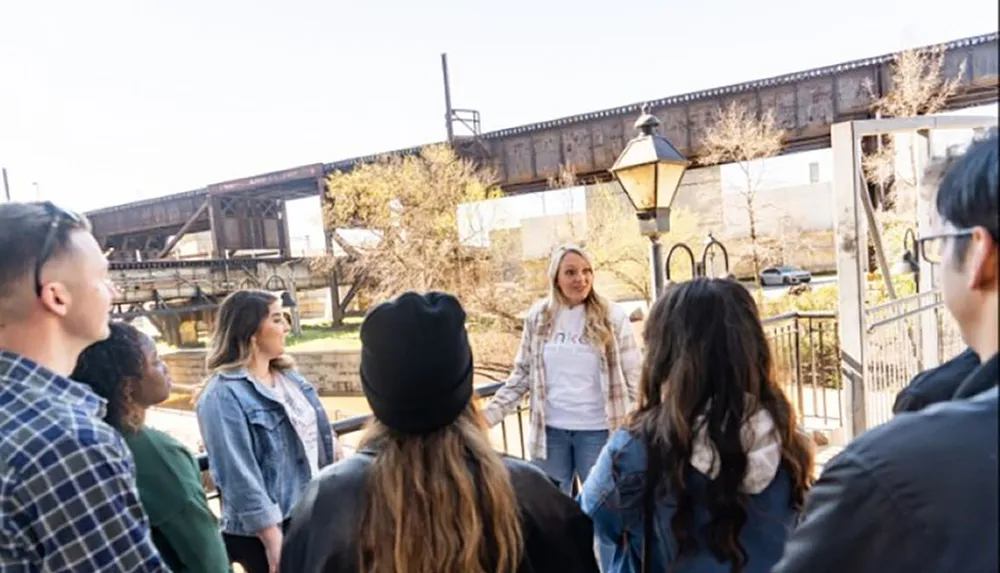 A group of people listens to a woman speaking possibly a guide at an outdoor location with a bridge structure in the background