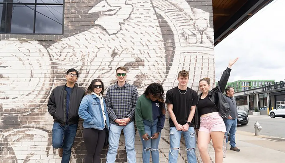 A group of young people are standing in front of a brick wall with a large mural with one person pointing upwards toward the artwork