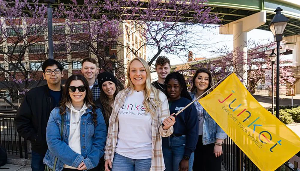 A group of cheerful young adults holds a Junket flag while posing together outdoors with blooming purple flowers in the background