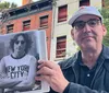 A person is posing with an open book that shows a photo of an individual wearing a New York City shirt seemingly standing in front of the same building that appears in the background