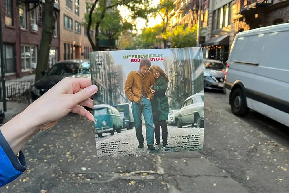 A person is holding up the album cover The Freewheelin Bob Dylan to seamlessly match the scene behind it which is the same location where the original album photo was taken