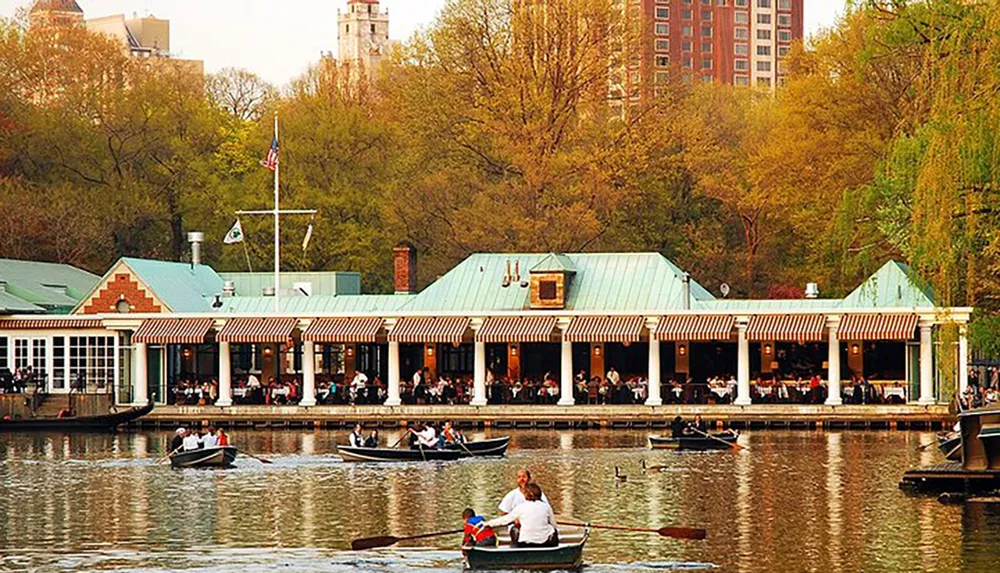 The image depicts a bustling lakeside restaurant with a striped awning where patrons are dining al fresco complemented by people leisurely rowing boats on the water in the foreground all set against a backdrop of lush greenery and city architecture