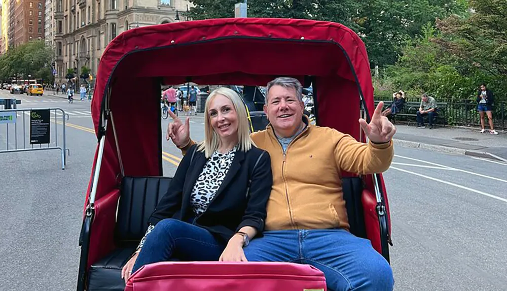 Two people are smiling and making peace signs while sitting in a red pedicab on a city street likely enjoying a leisurely tour