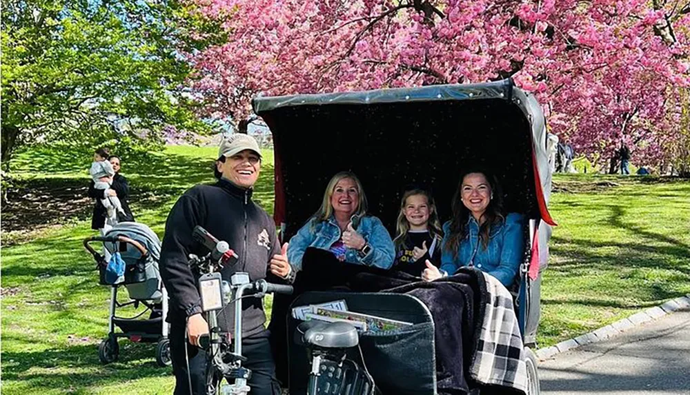 A group of people gives thumbs up as they pose for a photo in a park with blooming pink cherry blossoms with a smiling pedicab driver in the foreground