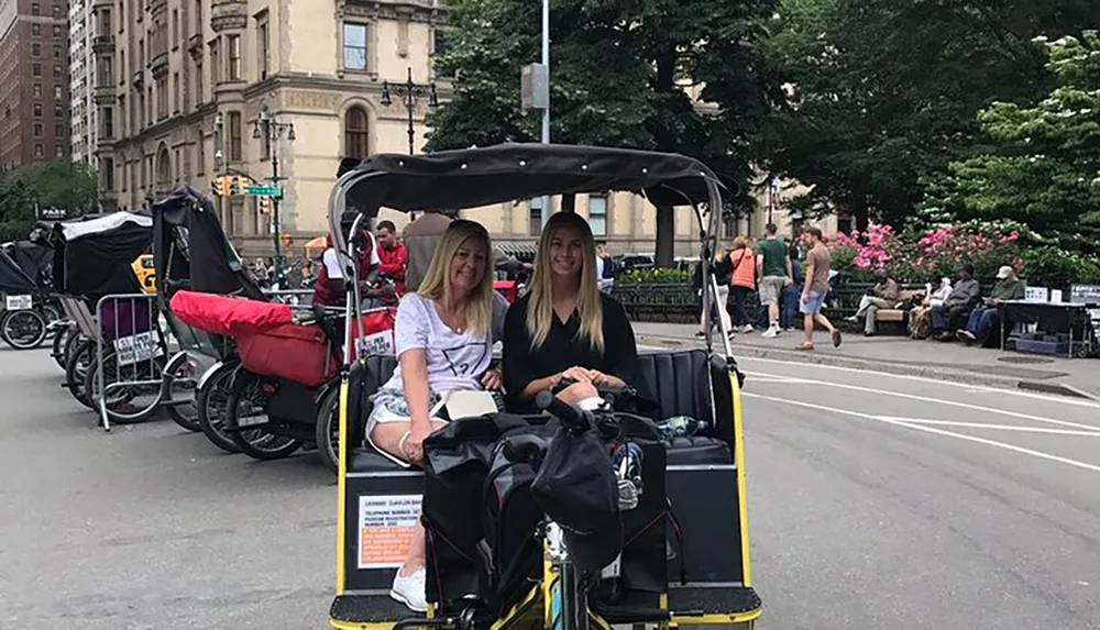 Two smiling people are sitting in a pedicab on a city street with other pedicabs and pedestrians in the background